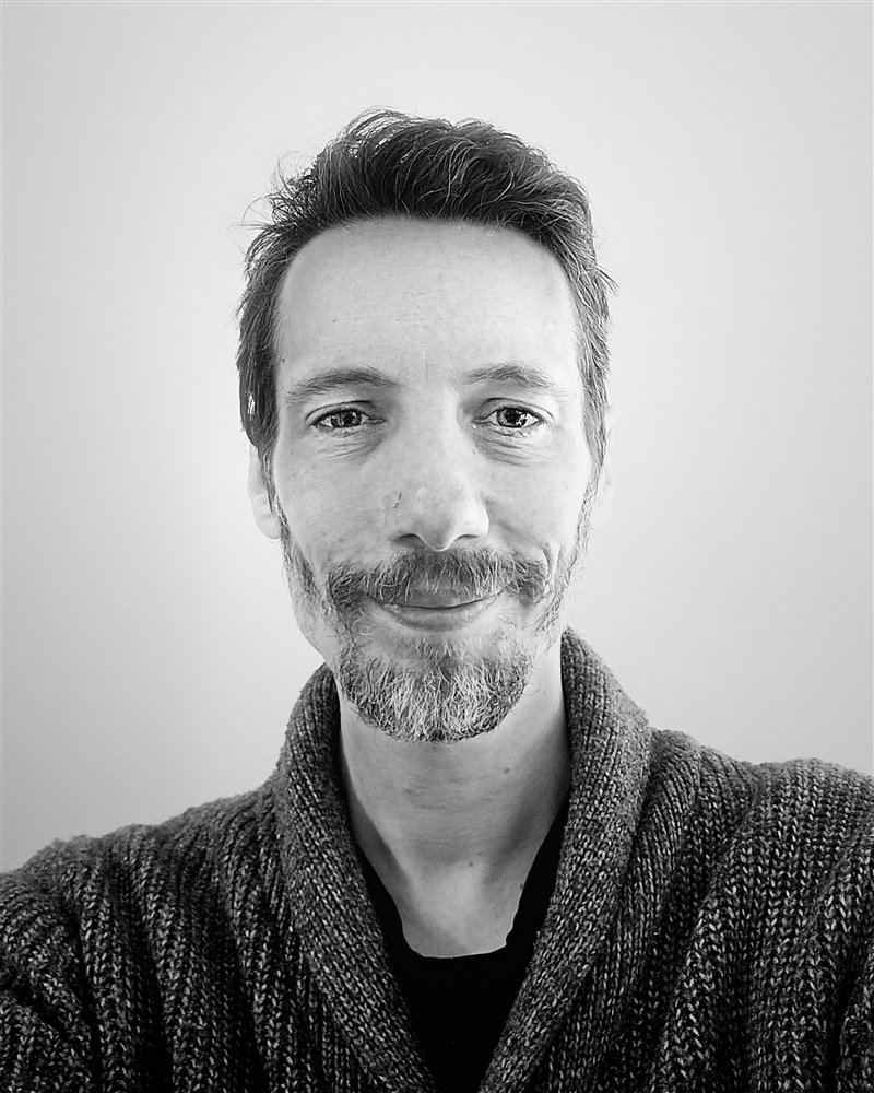 Black and white portrait of Paul London, smiling, in a knitted cardigan.