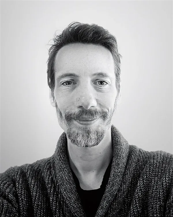 Black and white portrait of Paul London, smiling, in a knitted cardigan.
